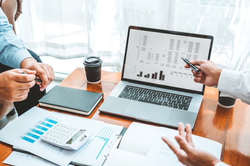Mãos de dois gestores conversando sobre a avaliação de desempenho financeiro da empresa, mostrada no laptop e em gráficos impressos sobre a mesa.