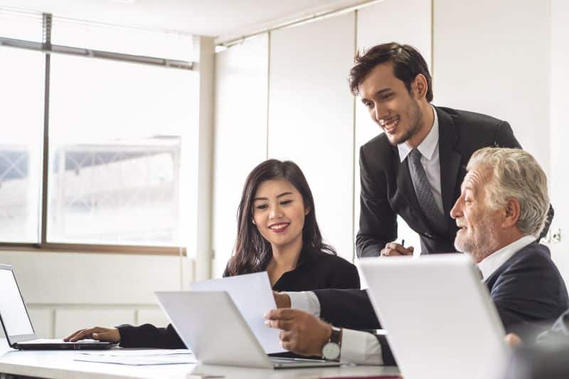 Uma mulher e dois homens, todos vestidos formalmente, em sala de reunião com computadores, analisam contrato SaaS e conversam sobre sua elaboração.