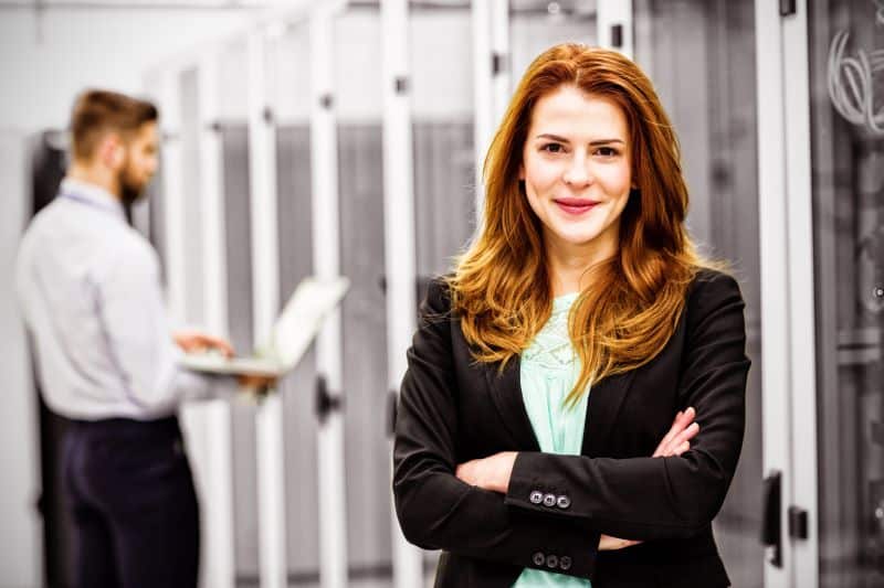 Mulher de braços cruzados em sala de servidor com olhar simpático para a câmera, com colega trabalhando ao fundo, satisfeita com a adoção de plano recorrente em sua empresa.