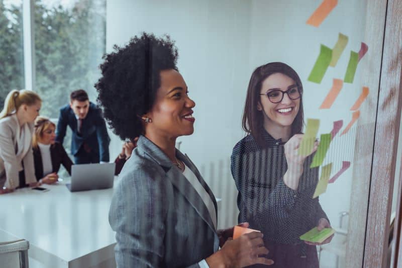 Duas mulheres falando sobre previsão de faturamento, utilizando post-its na divisória de vidro, com colegas ao fundo reunidos trabalhando.
