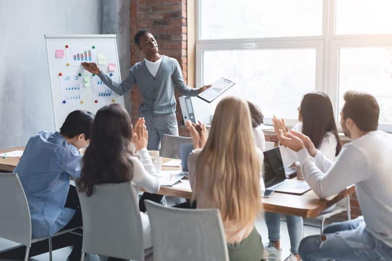 Homem em sala de reunião explica em pé aos seus colegas sobre o mapeamento de mercado, apontando informações em quadro branco.