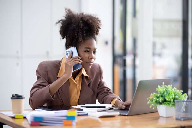 Gestora de marketing falando ao celular enquanto analisa ativos financeiros no computador.