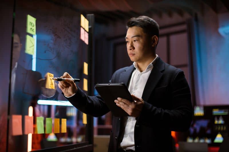 Gestor de TI calculando a relação entre ativo circulante e não circulante que tem em sua empresa com a ajuda de post-its e um tablet.