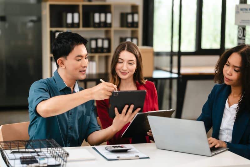 Empresário mostrando balanço financeiro no tablet para outras duas gestoras contábeis que analisam notebook e relatórios.