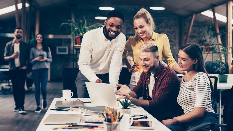 Como funcionam as startups: grupo de mulheres e homens sorridentes reunidos em volta de mesa com computador.