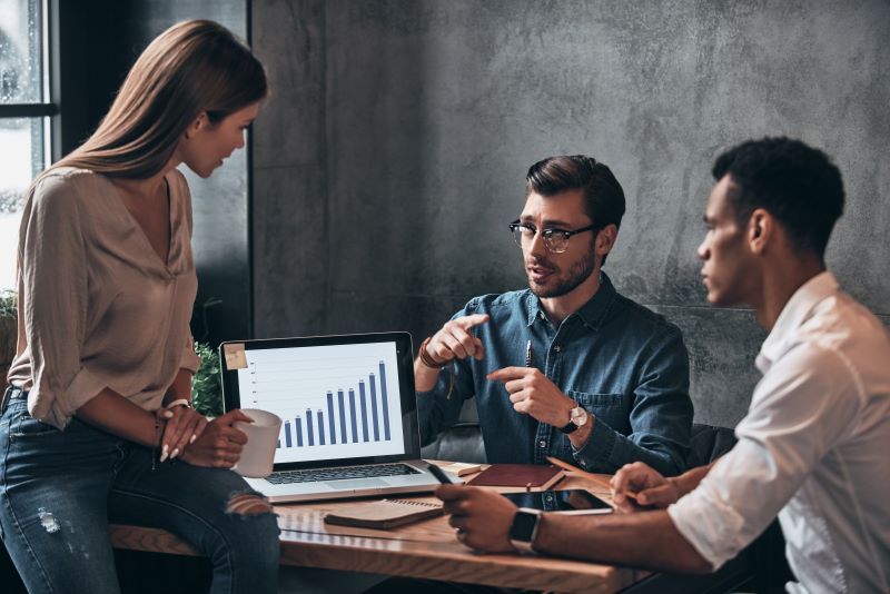 Dashboard o que é​: três profissionais reunidos em volta de mesa analisando gráfico no computador.