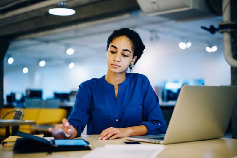 índices de liquidez​: mulher trabalhando em escritório fazendo contas em celular ao lado do notebook